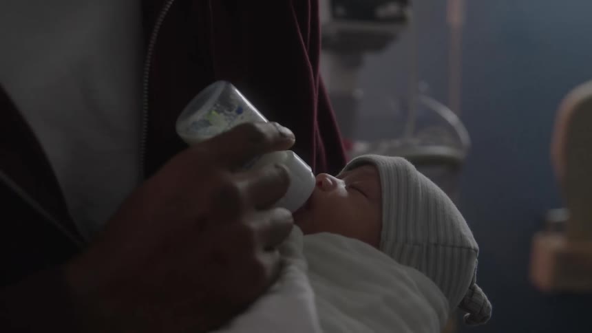 An adult holding an infant wrapped in a blanket and wearing a hat while placing a bottle to the babys mouth