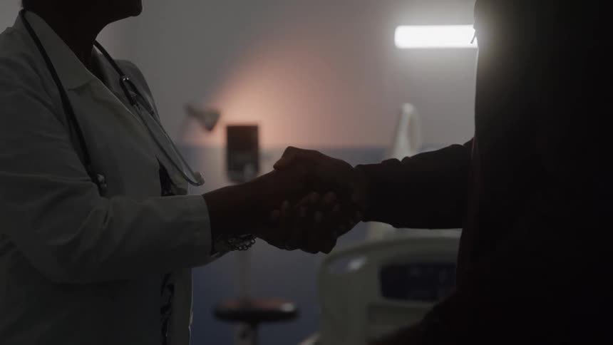 A doctor and man shake hands in a hospital in front of a bedridden patient