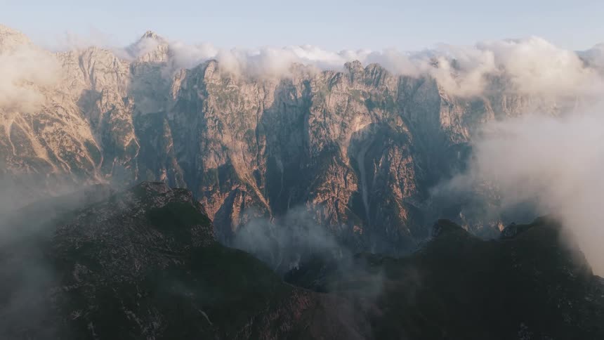 An aerial wide view shows dramatic rocky mountain cliffs shrouded in moving white clouds and mist under a clear evening sky creating a mystical atmosphere