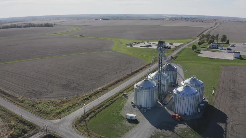 A farm storage facility with multiple grain silos stands amid a wide expanse of farmland at midday