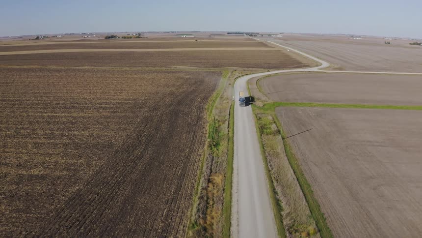 Blue truck with yellow trailer travels along a narrow road between agricultural fields