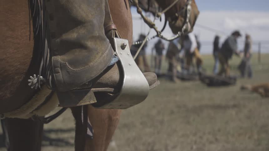 A closeup view focuses on a cowboy boot with a spur resting in a silver metal stirrup attached to a brown leather saddle on a horse with blurred ranch workers in the background