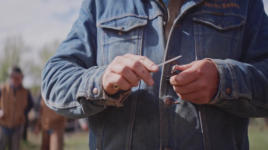 A closeup view focuses on a cowboys hands wearing a denim jacket as he expertly twists a piece of wire with a tool in the daylight with blurred people in the background