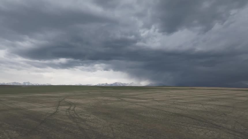 A wide expanse of open land transitions from brown to green under a layer of dark overhead clouds as precipitation falls in the distance towards a mountain range