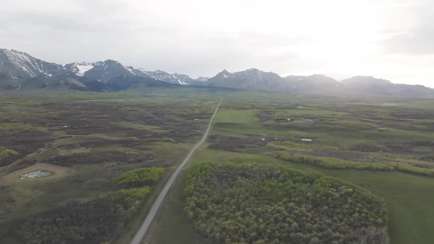 An aerial view rises over a long rural road passing through green fields and patches of woodland towards a range of snowcapped mountains under a bright overcast sky
