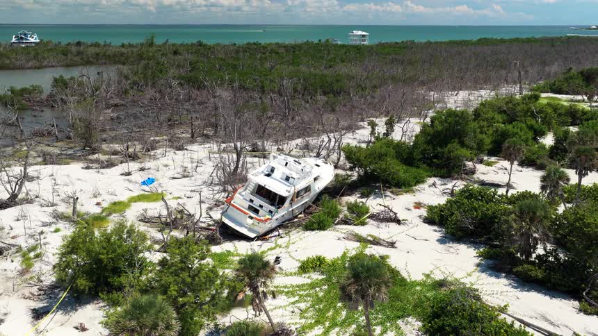 A white motor yacht sits beached and tilted on bright sand among scrub and dead trees with turquoise water and a few boats visible in the distance