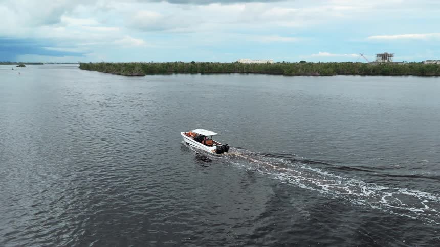 An aerial shot tracks a white speedboat with several people on board as it cruises along a dark river past lush tree lined shores