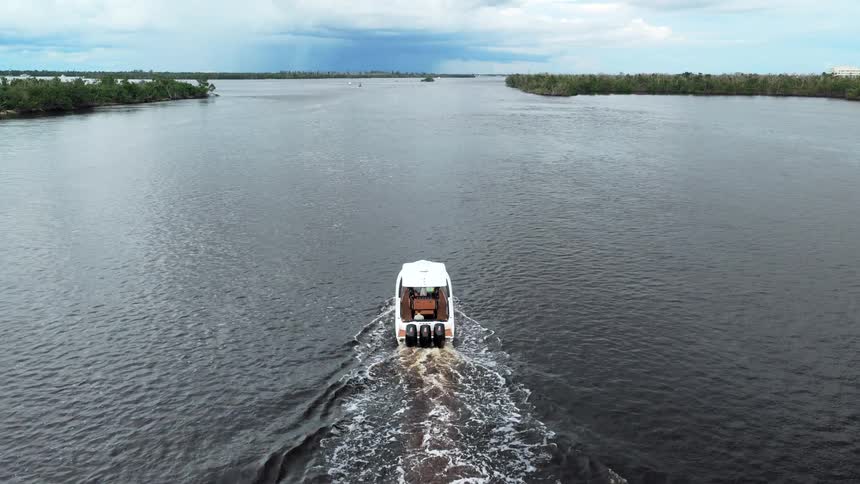 A white motorboat with three outboard engines moves across a wide dark river leaving a foam wake as it travels between two treelined banks under a cloudy sky