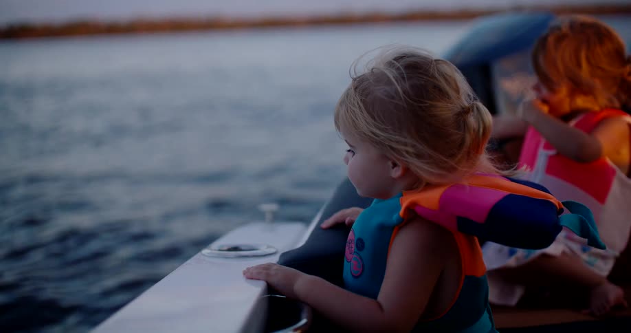 A young child in a life jacket looks out intently over the water from the side of a boat at dusk