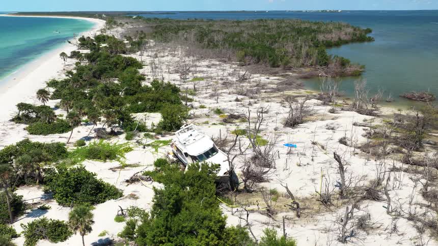An aerial wide view shows a white yacht severely beached on a sandy shoreline surrounded by dead trees and green shrubs located near a bay and a white sand beach under a clear blue sky