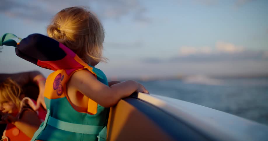 Close up of a female toddler wearing a safety jacket and standing in a moving boat during daytime