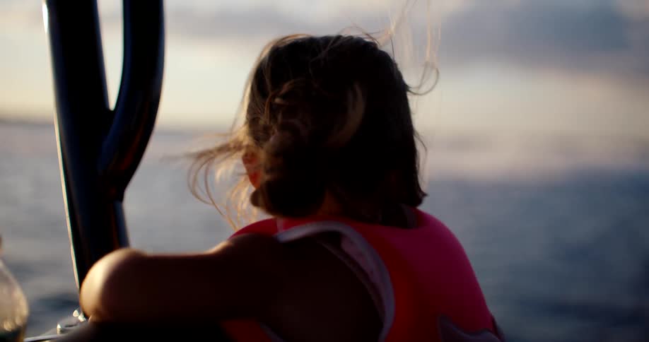 Child wearing a life jacket watches the sea at sunset leaning on the side of the boat