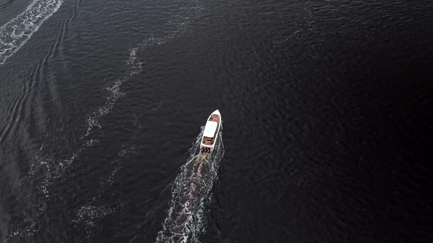 An overhead drone view tracks a small white motorboat speeding across dark water leaving a white foamy wake trailing behind it