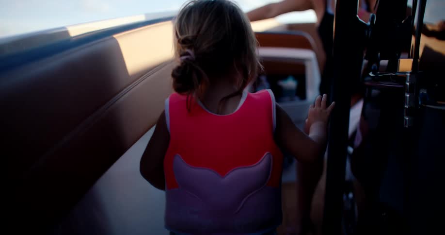A young girl with blonde hair in a pink and orange life jacket stands with her back to the camera on a boat deck with an adult visible in the background