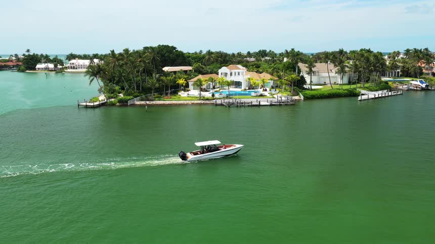 An aerial wide view shows a white speedboat moving across bright green canal water past a large luxury waterfront home with a pool surrounded by palm trees on a sunny day