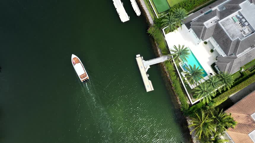 House with white roof and pool surrounded by palm trees by canal with boat approaching