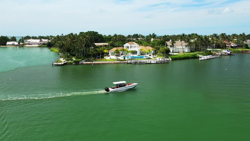 A speed boat cruises through vibrant green water past luxurious waterfront homes and lush palm trees under a bright sky