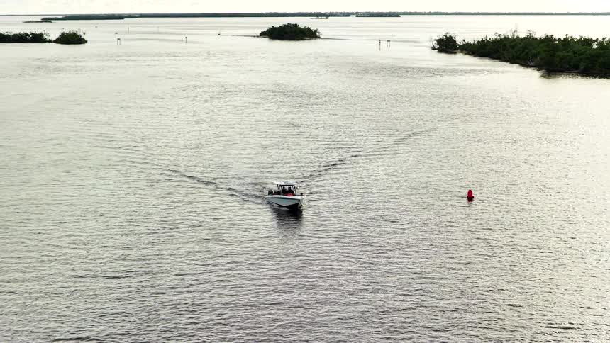 An aerial view tracks a small white speedboat cruising on calm graygreen water past a red channel marker and small mangrovecovered islands under a bright overcast sky