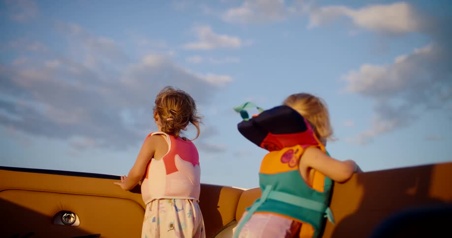 Two young girls with blonde hair wear bright life jackets and stand looking out from the brown vinyl seats of a boat against a blue sky with white clouds