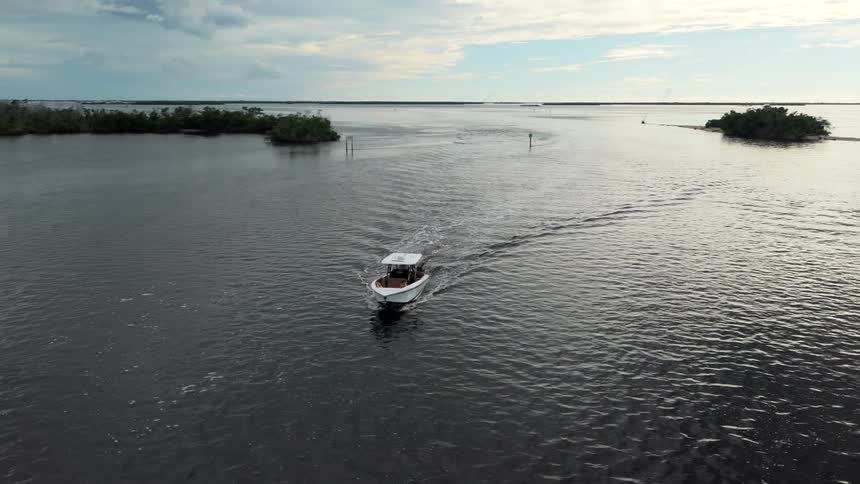 An aerial wide view tracks a white and brown small motorboat moving across dark water leaving a wake passing small mangrove islands and a channel marker under a cloudy sky