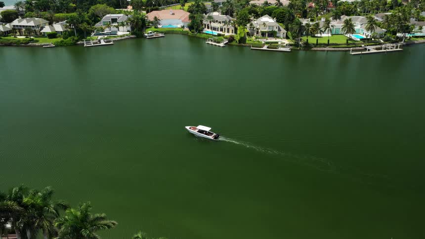 A small watercraft moving across a wide body of water flanked on both sides by a shoreline densely populated with houses pools and tropical foliage