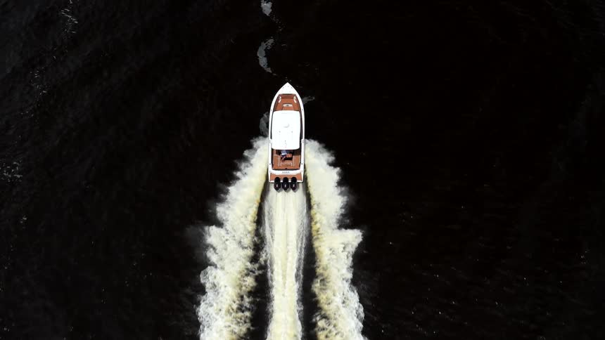 An overhead drone view follows a white motorboat with triple outboard engines as it moves fast through dark water leaving a long churning wake behind it