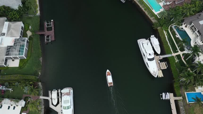 An overhead aerial view tracks a small brown and white motorboat moving down a dark green canal lined with docks and large luxury homes with docked yachts on a bright day