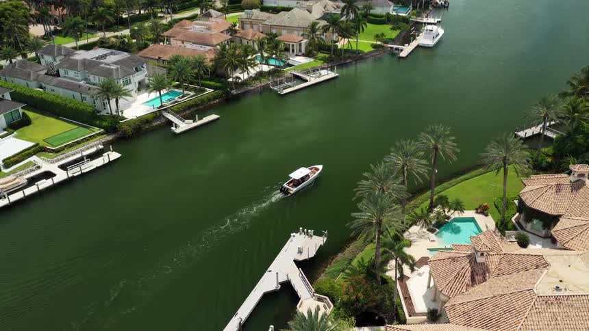 Boat sails slowly through a canal surrounded by houses with pools and palm trees