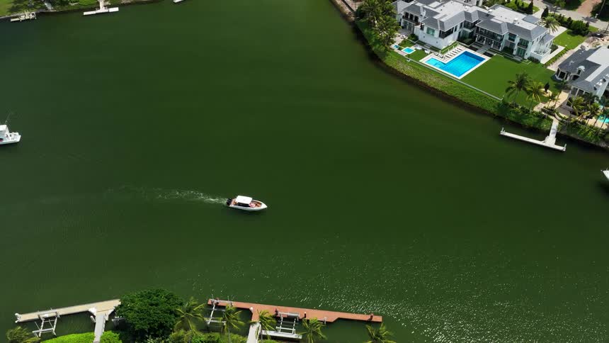 An overhead aerial view tracks a small white motorboat cruising across dark green canal water past luxurious waterfront homes with pools and private docks on a sunny day