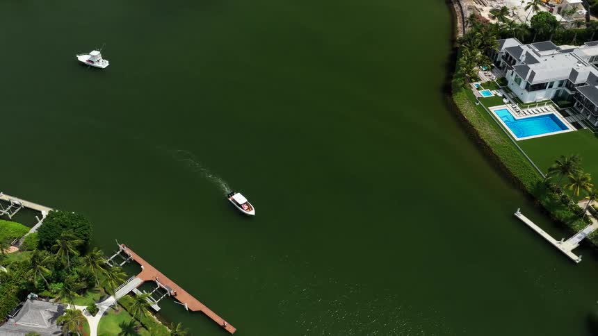 An overhead aerial view tracks a small white motorboat cruising across a dark green canal past luxurious waterfront homes with blue swimming pools and private docks on a sunny day