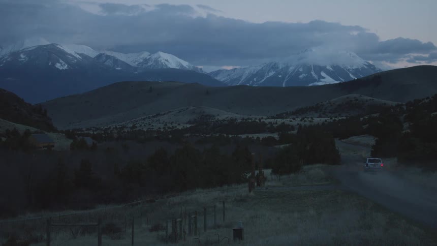 Car drives along a rural road at dusk with snowcapped mountains in the background