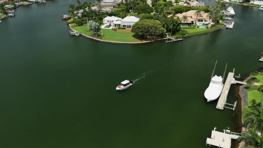 Aerial view of a calm greenish canal lined with upscale homes and docks a small boat cruises past a moored yacht toward a lush palmstudded island yard