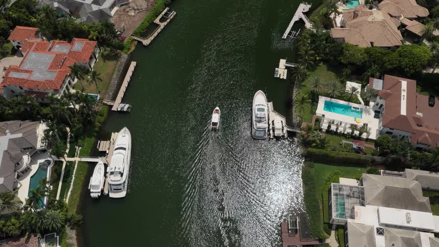An overhead aerial view tracks a small motorboat moving slowly down a dark green canal flanked by luxurious waterfront homes docks and several large docked yachts on a bright sunny day