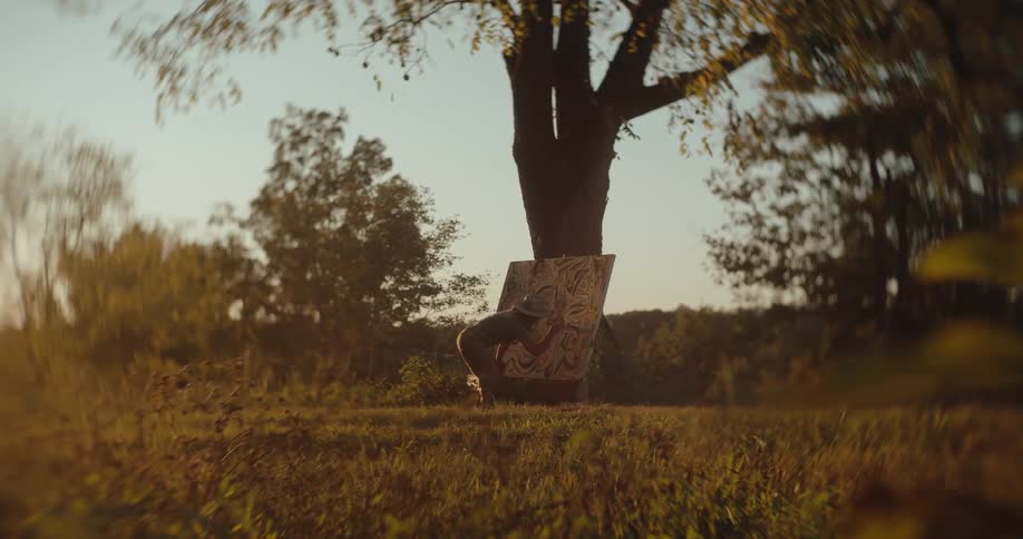A wide lowangle view shows an artist leaning against a large tree trunk painting on a tall canvas in a grassy field at sunset creating a warm golden glow