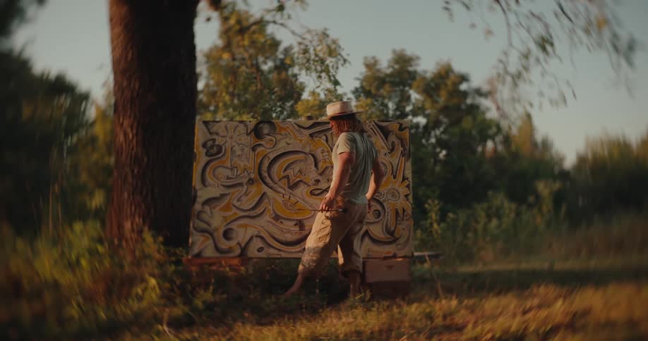 An artist wearing a hat and light clothes stands in a grassy field at sunset working on a large abstract painting attached to a wide tree trunk