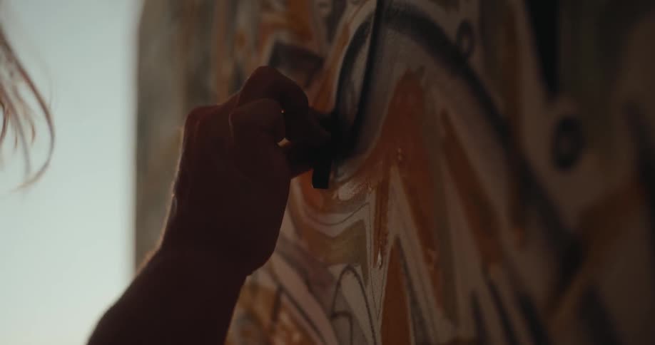 A close view shows an artists hand wearing a dark glove as they apply thick paint to a canvas featuring an abstract design with warm orange and brown colors