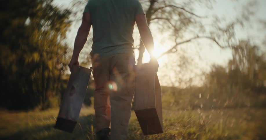 Man carrying two wooden frames walks through a field near the woods on a sunny day