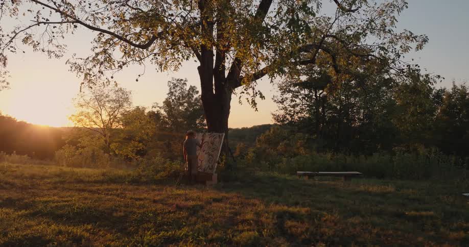 Person painting outdoors at sunset in front of a tree with canvas propped up