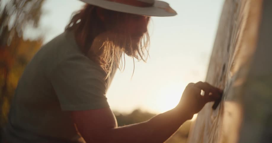 A closeup side view shows a person with long hair wearing a cowboy hat and a tshirt drawing on a large outdoor canvas with warm evening sun flare washing out the background