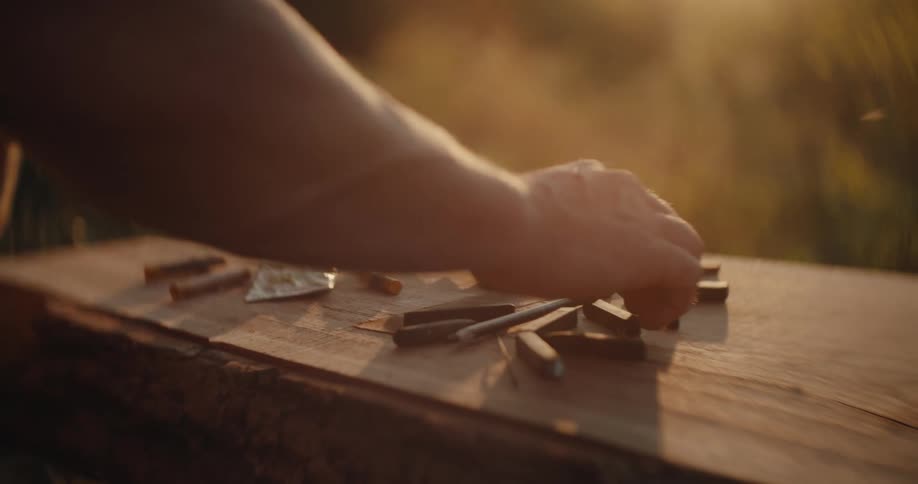A closeup view shows an artists hands picking up and handling charcoal sticks and a pencil resting on a wooden outdoor table in warm golden sunset light