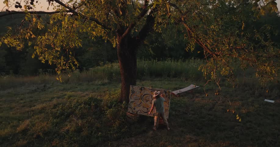 An elevated wide view shows an artist wearing a hat and light clothes painting a large abstract canvas attached to a wide tree trunk in a grassy golden field at sunset