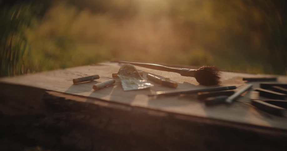 A wide close view shows a collection of charcoal sticks brushes and art tools scattered on a wooden outdoor table with golden sunlight shining through the blurred green background