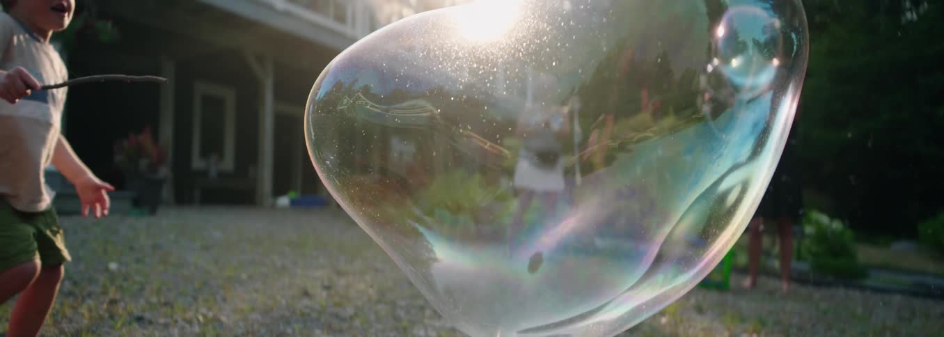 A large soap bubble floats and bursts in the sunny backyard of a house with a young child and adults visible their reflections shimmering on the bubbles surface