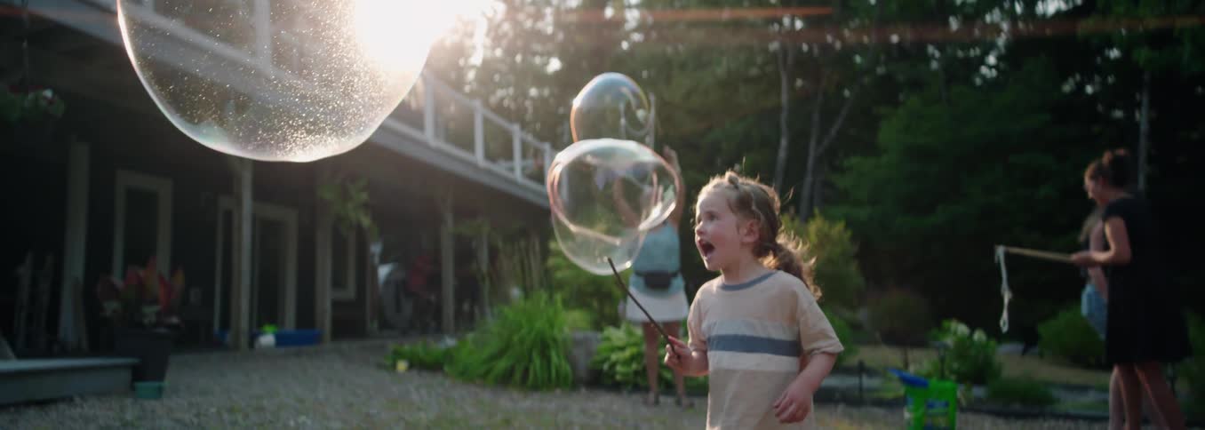 Girl pops a large bubble while she and her family play with bubbles in the yard of a rural residential building