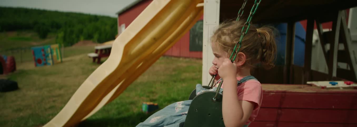 Little girl in pink shirt and blue overalls sitting on playground swing