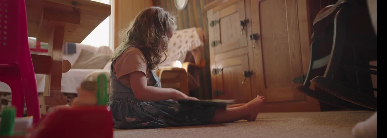 A young girl with long curly blonde hair sits barefoot on a light carpet in a sunlit living room reading a picture book against a background of wooden furniture