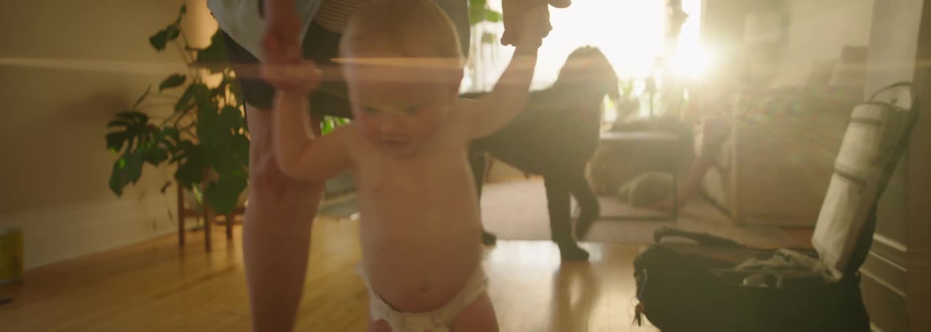 A smiling baby wearing only a diaper is held by the hands and takes assisted steps on a wooden floor in a sunlit living room with a black dog in the background