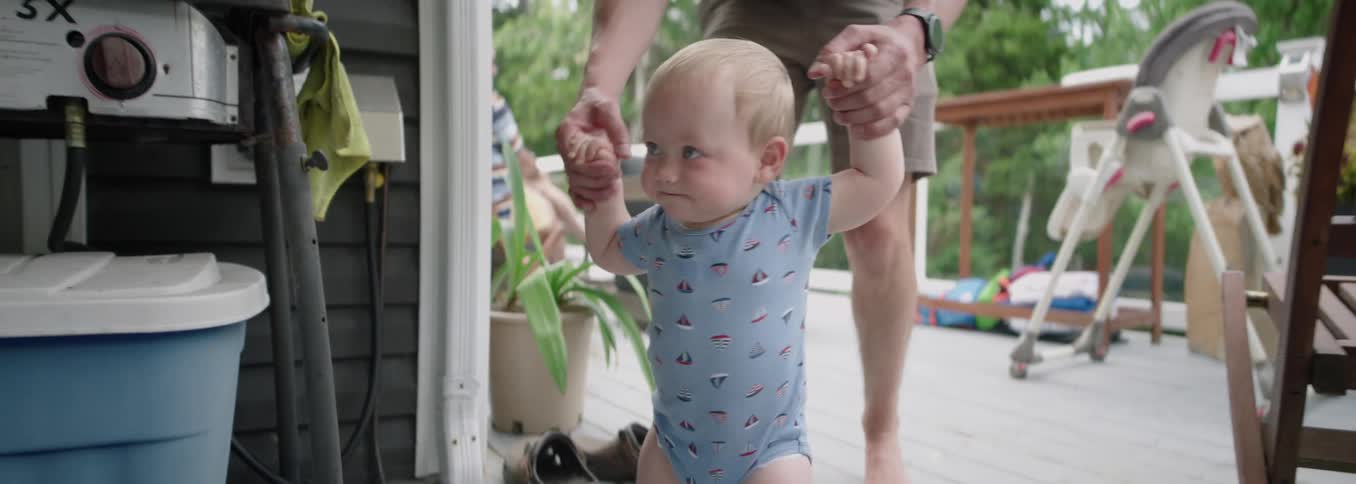 Baby takes steps with the support of an adult in an outdoor setting
