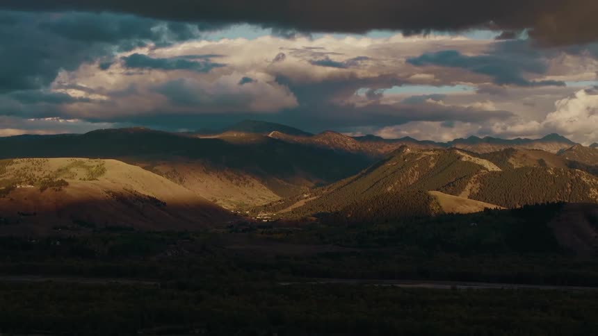 An establishing wide view shows a dramatic shadowed valley nestled between rocky mountains with a patch of sunlight highlighting a peak under a cloudy evening sky