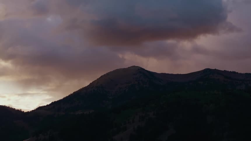 A mountain ridge at dusk under heavy purplegray clouds warm light glows along the horizon while the slopes fade into dark greens and browns